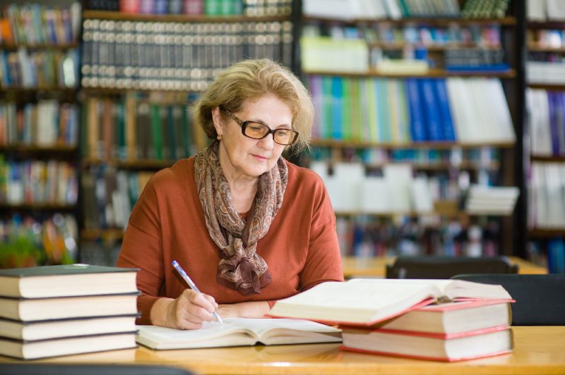 Senior woman sits in library surrounded by books at a table. She is writing in a book and also looking at one open in front of her.