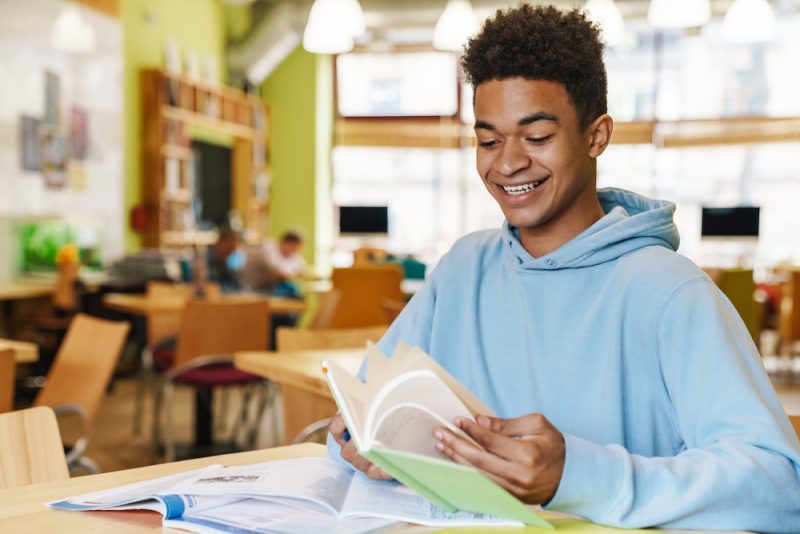 A smiling teenage boy sits at a desk in a coffee shop reading a book. He appears to be studying since there are more books open on the desk.