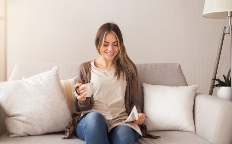Smiling woman sitting on couch and reading a magazine with a coffee mug in her other hand.