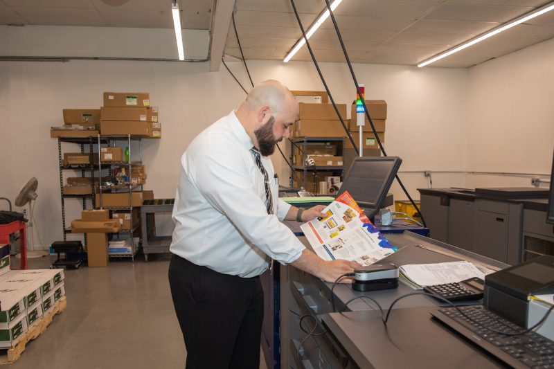 Austin Sullivan stands in the middle of our digital press room. He is examining one of our six-page stringer booklets printed from the digital press.