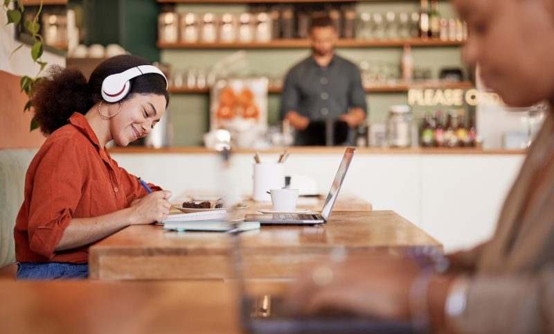 A young woman sits and works on her computer and writes in a notebook. She is in the middle of a coffee shop.
