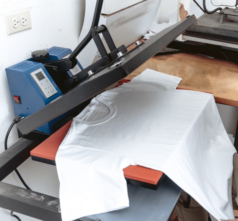 A white cotton t-shirt lays on a table with a transfer device poised above it. The device will lower down and place a design onto the t-shirt using heat.