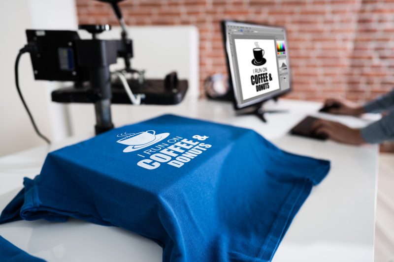 A machine uses heat transfer to place a design from a computer onto a t-shirt.