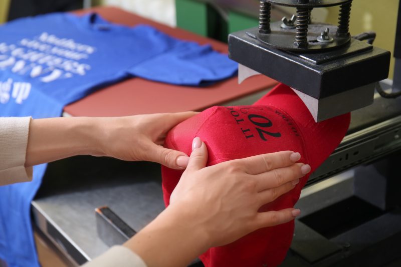A screen printing technique is used on a red baseball cap to transfer black text onto the hat.